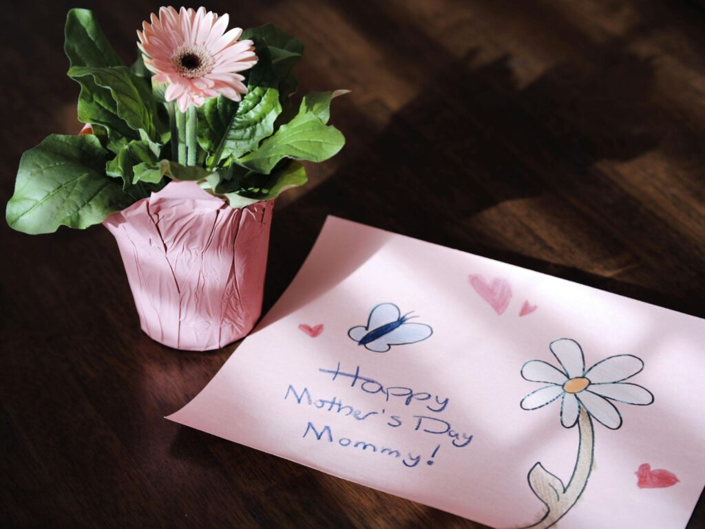 Una flor rosa en una maceta junto a una tarjeta hecha a mano que dice "¡Feliz Día de las Madres, mami!" sobre una mesa de madera.